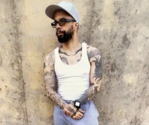 Man with tank top, tattooed arms, beard, sunglasses and a baseball cap