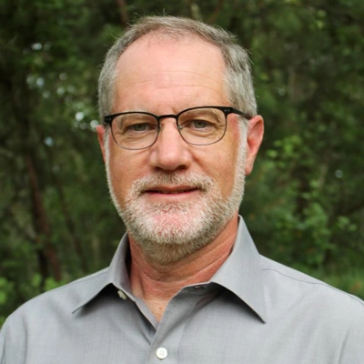 Caucasian man with gray hair and beard, glasses and collared shirt, thoughtful expression.
