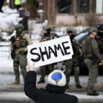 Person holding a sign saying SHAME with ICE troops in the background