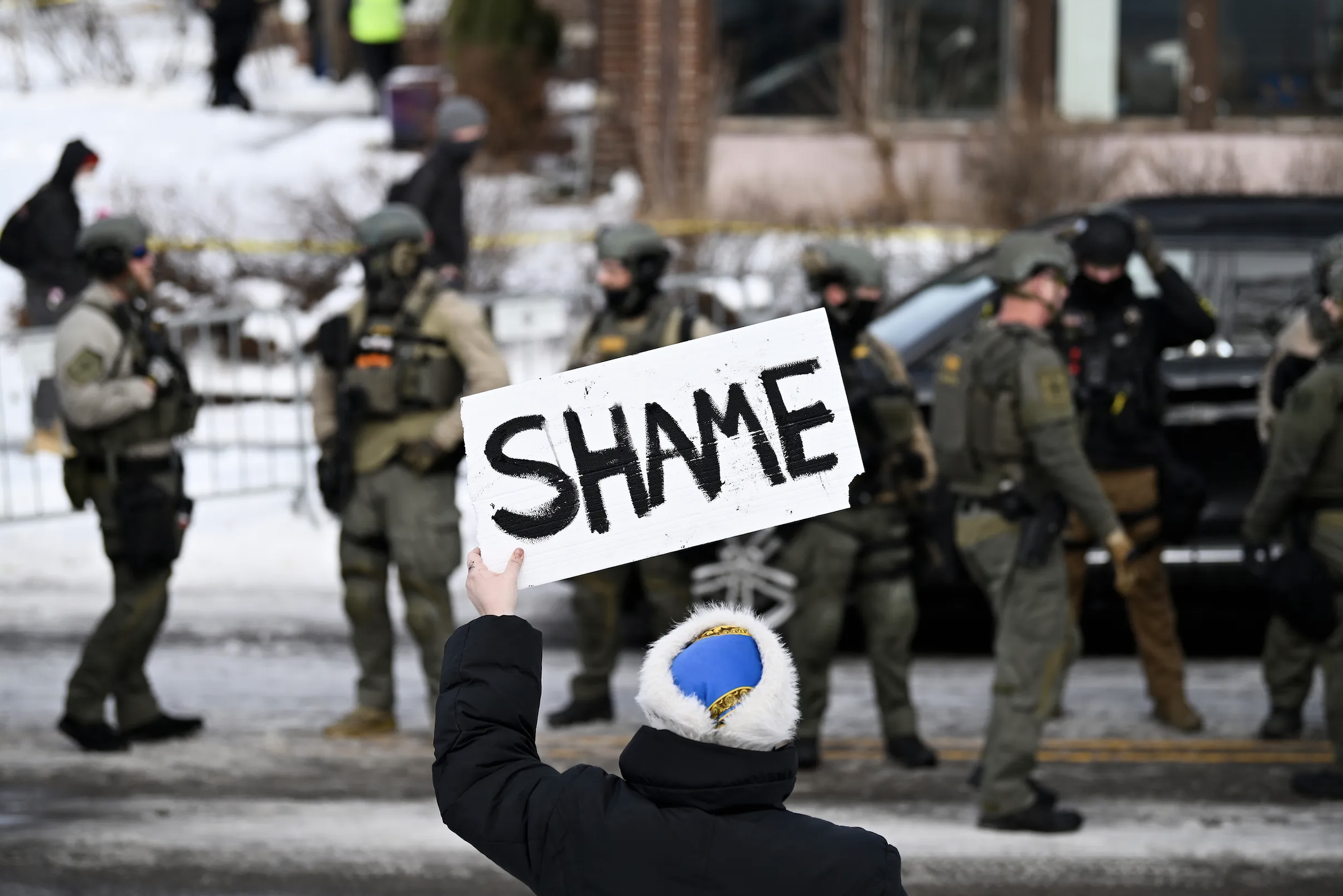 Person holding a sign saying SHAME with ICE troops in the background